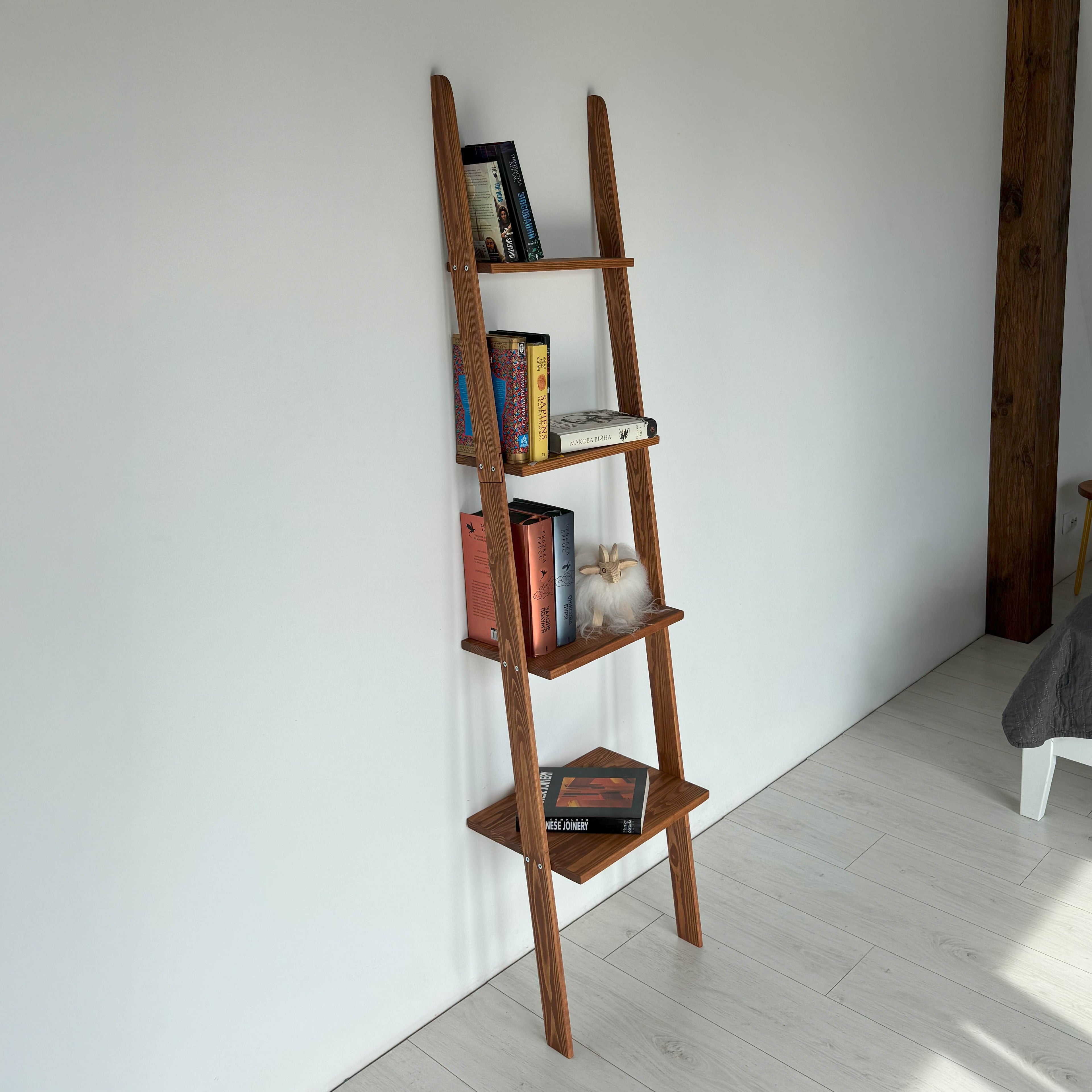 Wooden ladder shelf against a white wall with books on it, in a room with a bed and wooden floor.